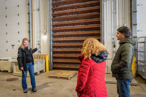A person gives a tour of a modern dry storage facility, gesturing to a floor-to-high-ceiling wall of crates.