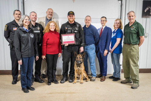 Ten people and one dog stand for a group photo. In the center, a police officer holds an plaque.
