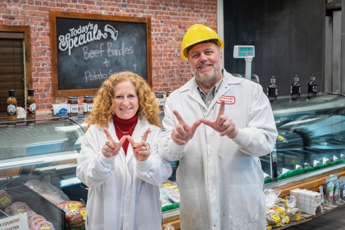 Two people wearing white coats stand in a deli and make a W sign with their hands while smiling to the camera.