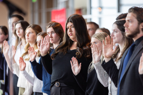 Students in an auditorium stand and hold up their right hands as they take a pledge.