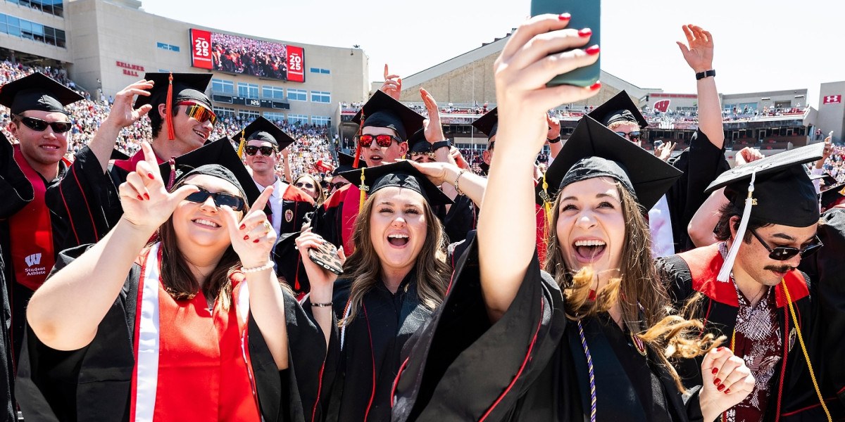 A group of graduates in black caps and gowns with red and white accents celebrate at a graduation ceremony, taking selfies and cheering in Camp Randall Stadium.