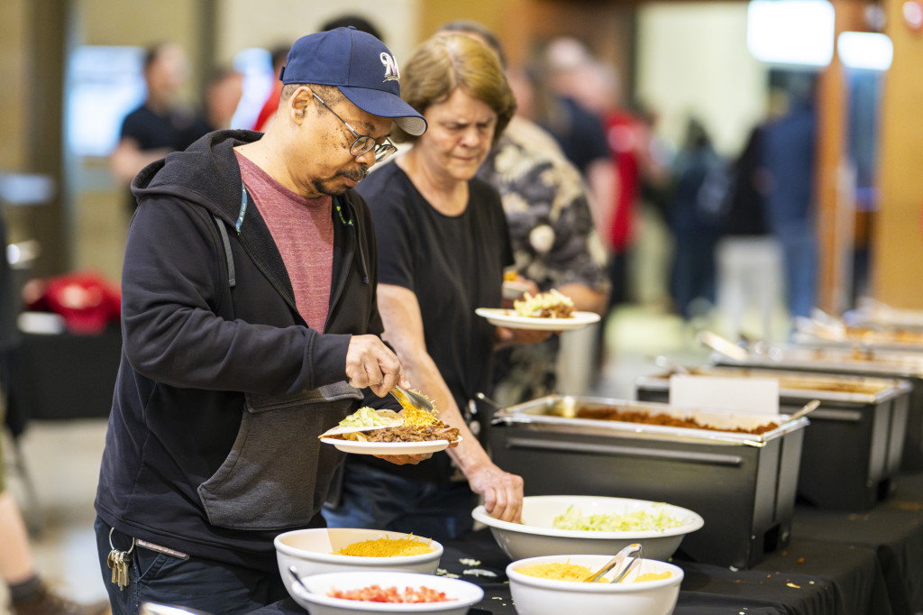 A man wearing a Milwaukee Brewers baseball hat stands in a buffet line. He is filling his plate with cheese that will top his tacos already plated on his plate.