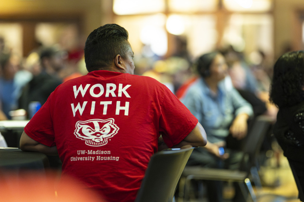 A employee wears a read shirt. His back faces the camera as he listens to a speaker who does not appear in the image frame. The back of the employee's shirt reads 'work with Bucky' and shows a picture of Bucky Badger.
