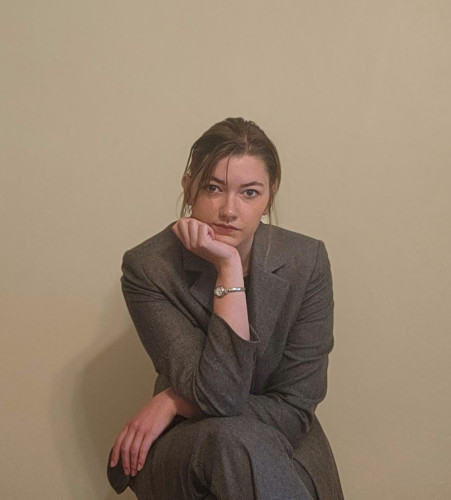 Grant sits on a chair in front of a white backdrop. She crosses her legs and her head rests on her hand that is propped up on her crossed leg.