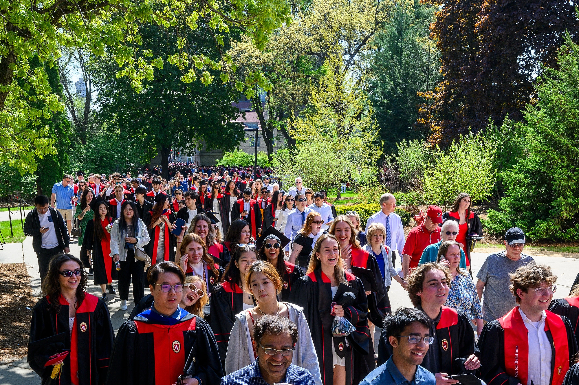 A crowd of people, some wearing graduation caps and gowns, walk along a wide path through a park setting on a bright, sunny day.