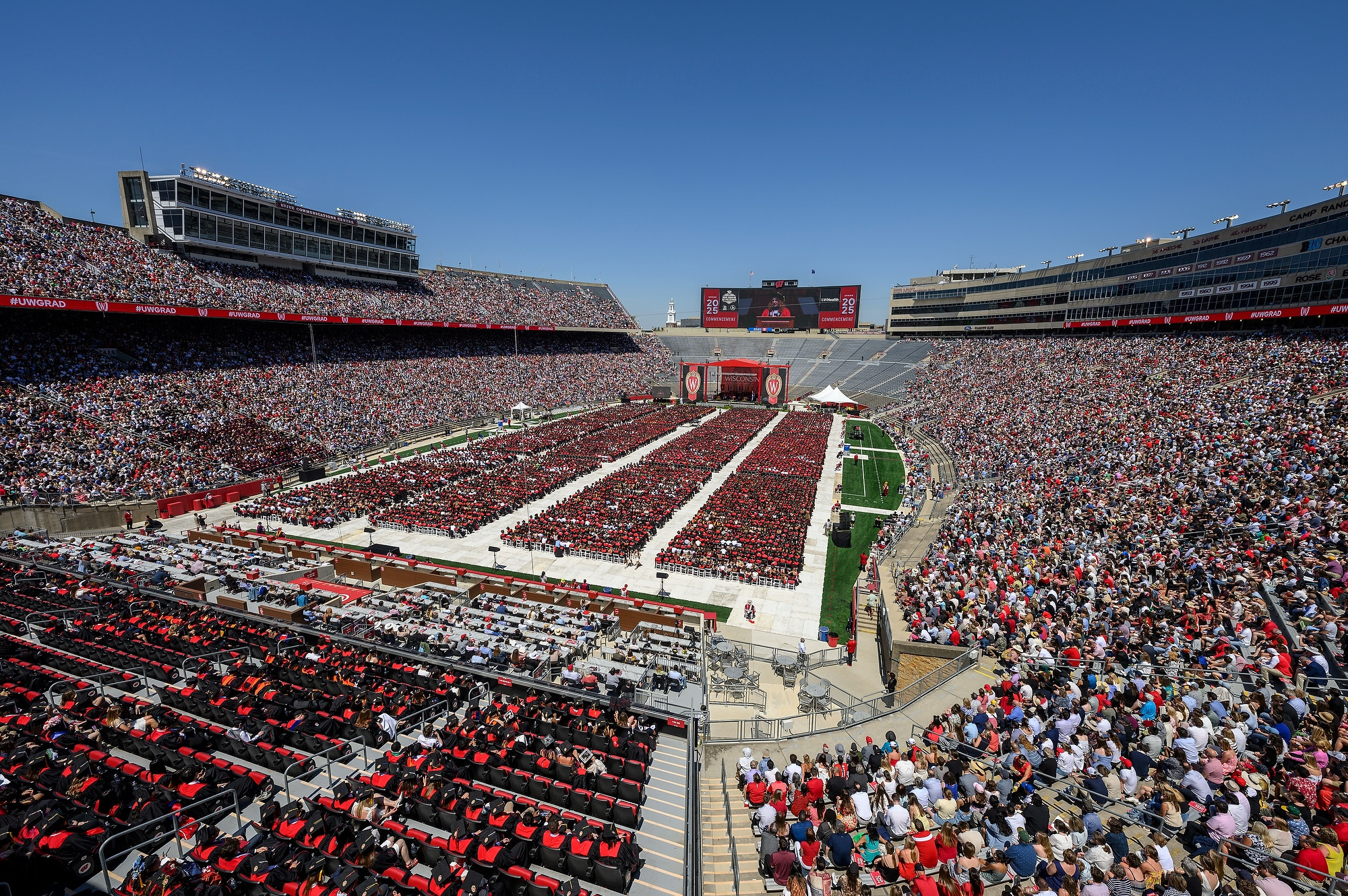 A crowd fills the stands at Camp Randall. On the field, graduating students in red and black gowns sit in packed rows of chairs facing a stage.