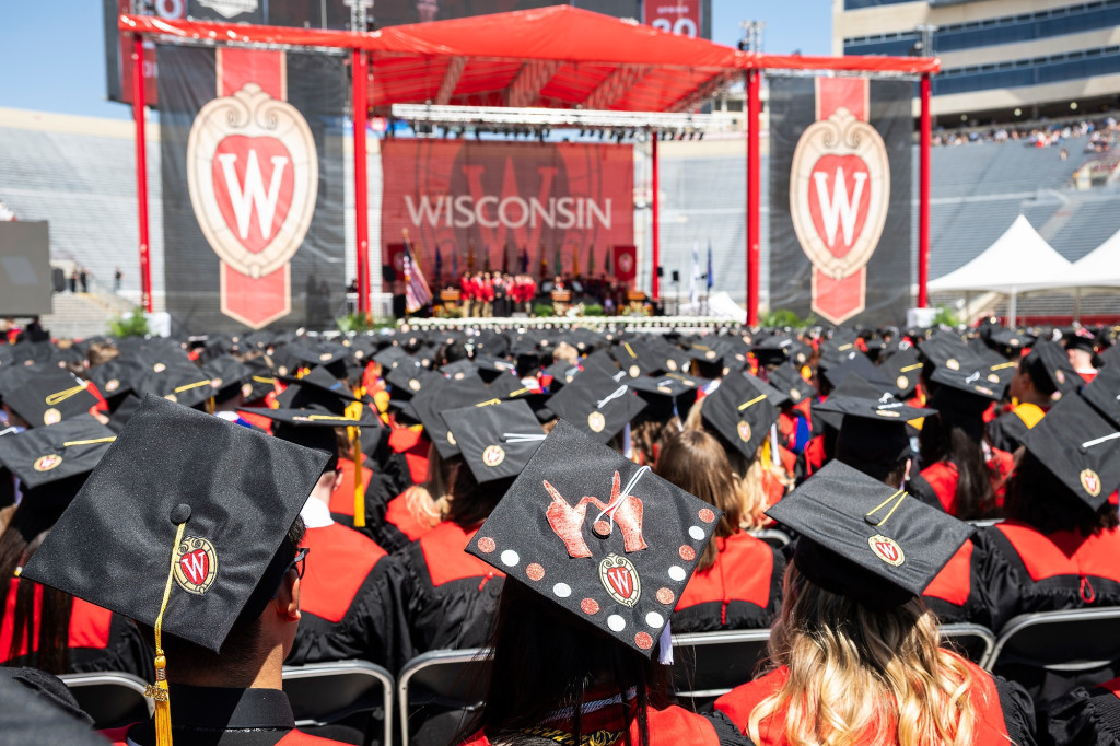 Graduates seated in a stadium look at a stage; their caps are visible from behind.