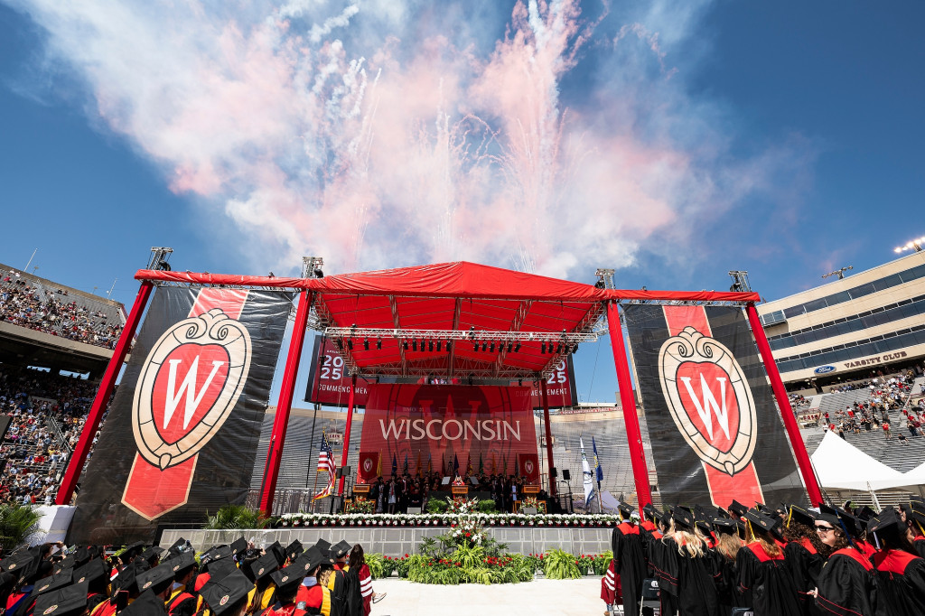 A stage decorated in red and white Wisconsin logos and staffed by academic leaders is shown, with fireworks going off overhead.