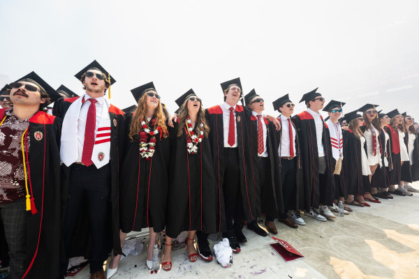 Graduates in cap and gown stand with their arms around each other and sing the university's alma mater.