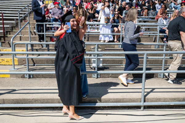A woman in cap and gown stands on the field and leans over a barrier to hug her sister in the crowd.