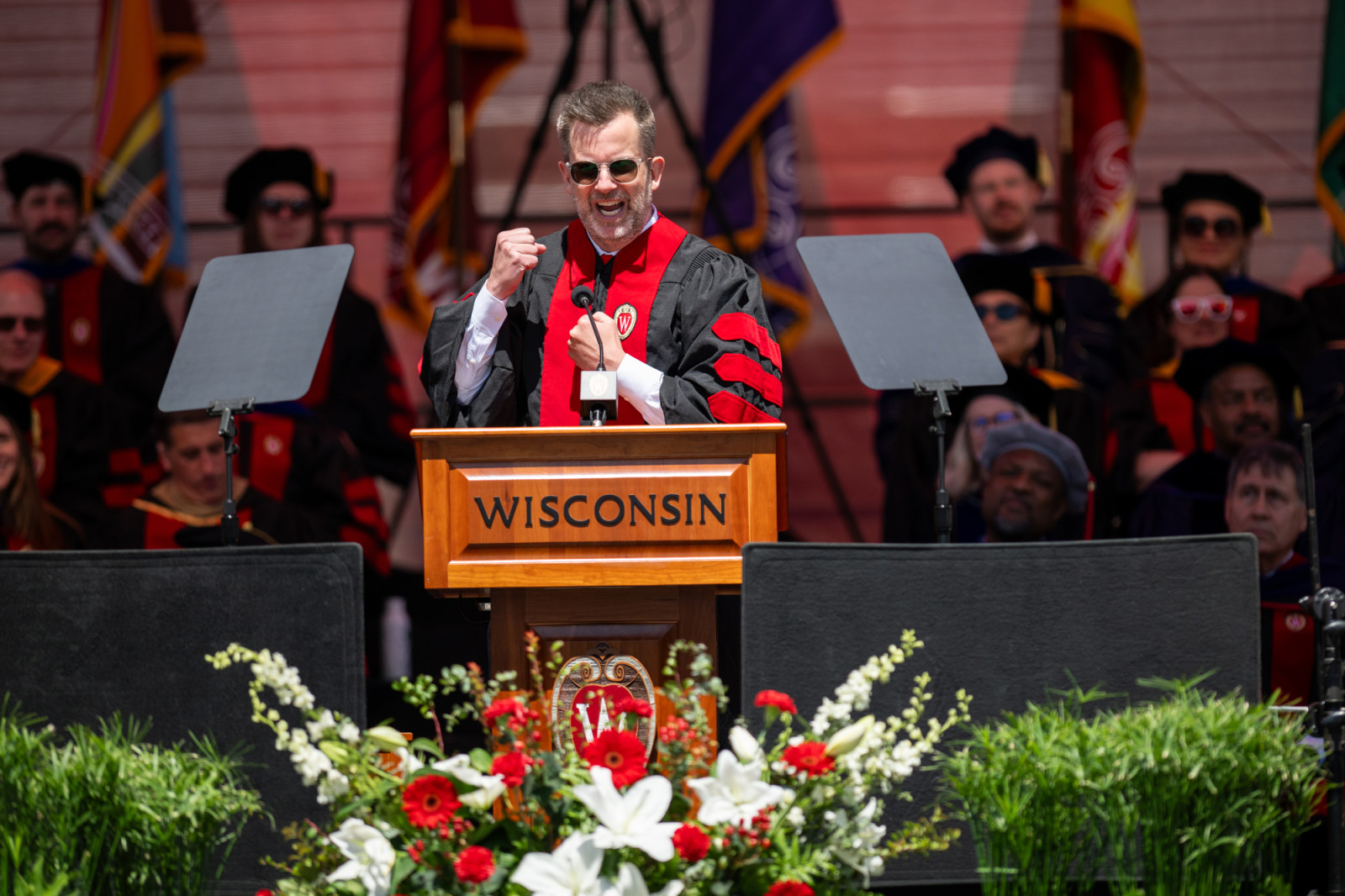 Jason Gay gestures and speaks while wearing a red and black academic robe and standing behind a podium.