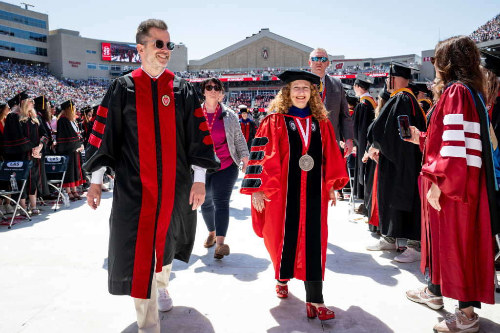 A number of people in academic robes walk down the aisle of a stadium.