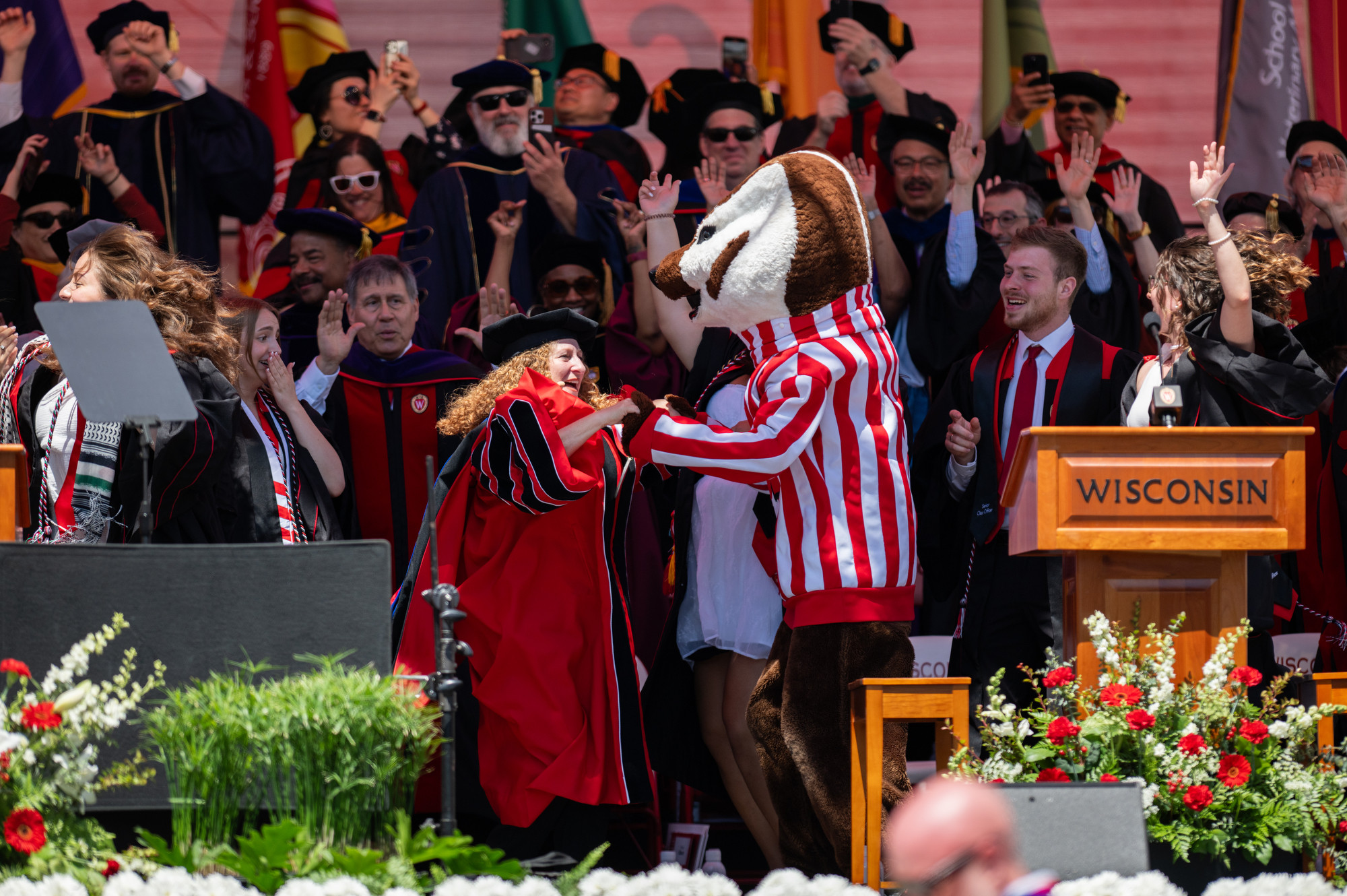 Chancellor Mnookin and Bucky Badger join hands and dance on stage as a crowd of people cheer and jump around them.