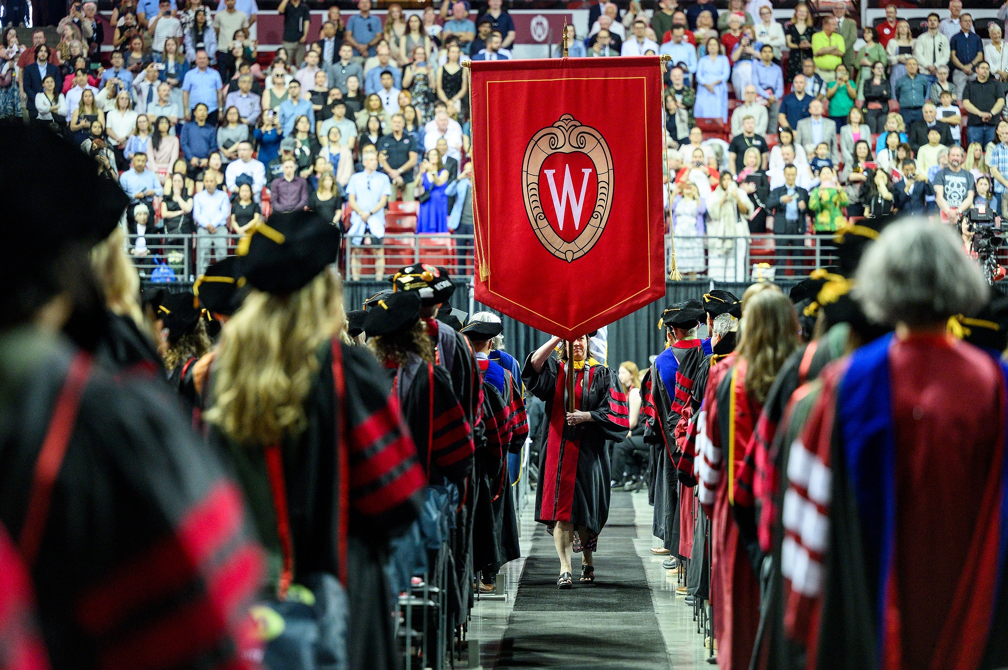 A graduate wearing cap and gown carries a red banner with the UW crest down a center aisle, surrounded by fellow graduates.