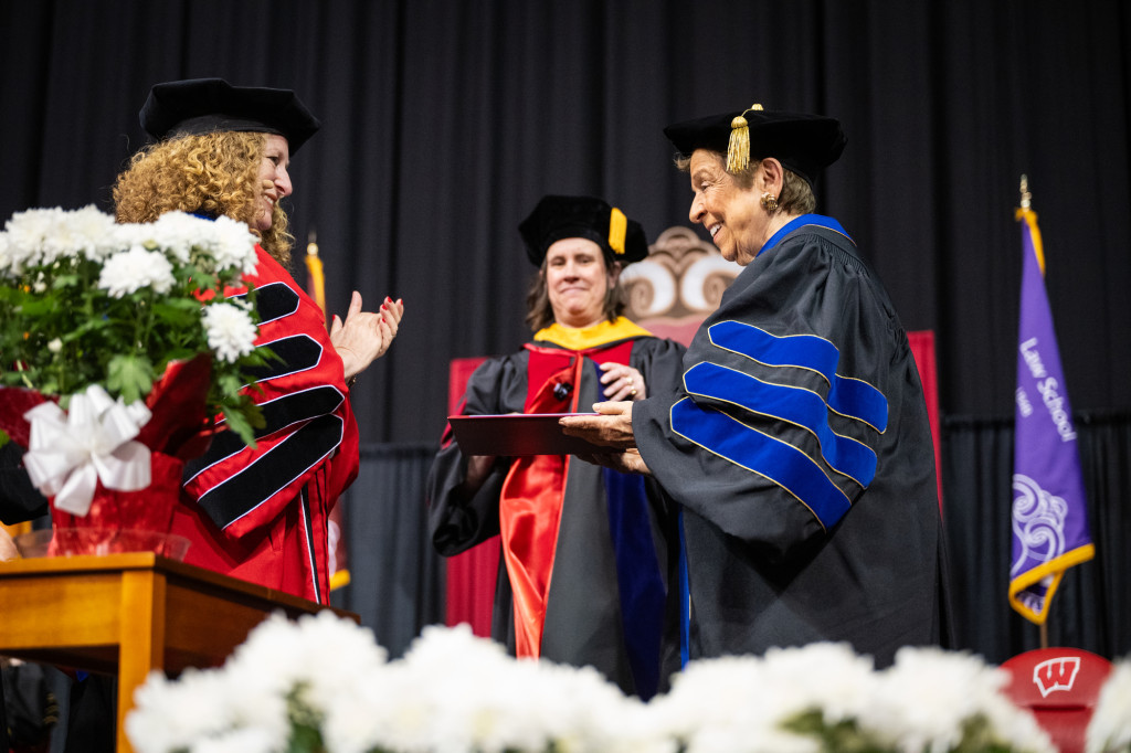 A woman gives a diploma to another woman, both wearing academic robes.