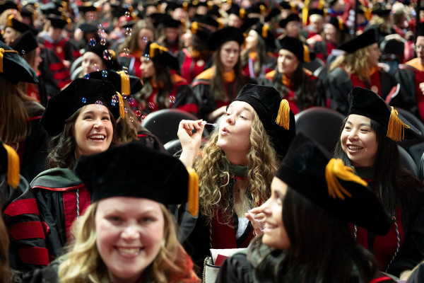 A woman blows bubbles while sitting among fellow graduates.