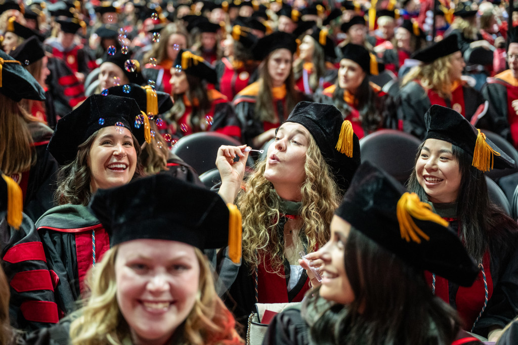 A woman in commencement robes, surrounded by other graduates, blows bubbles.