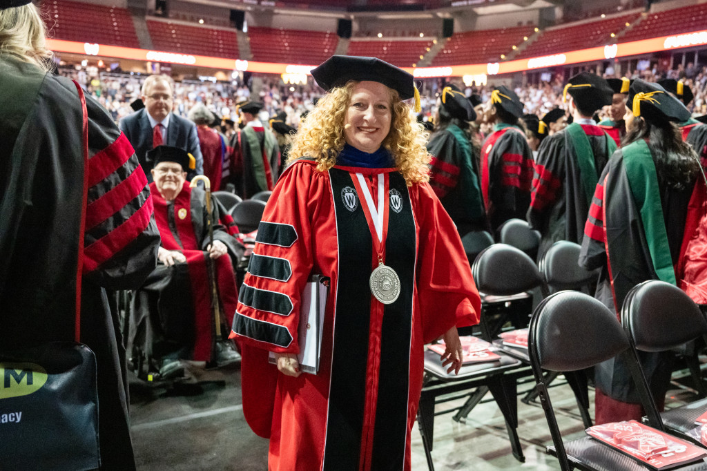 A woman in academic robes walks down an aisle, smiling, surrounded by others in commencement gowns.