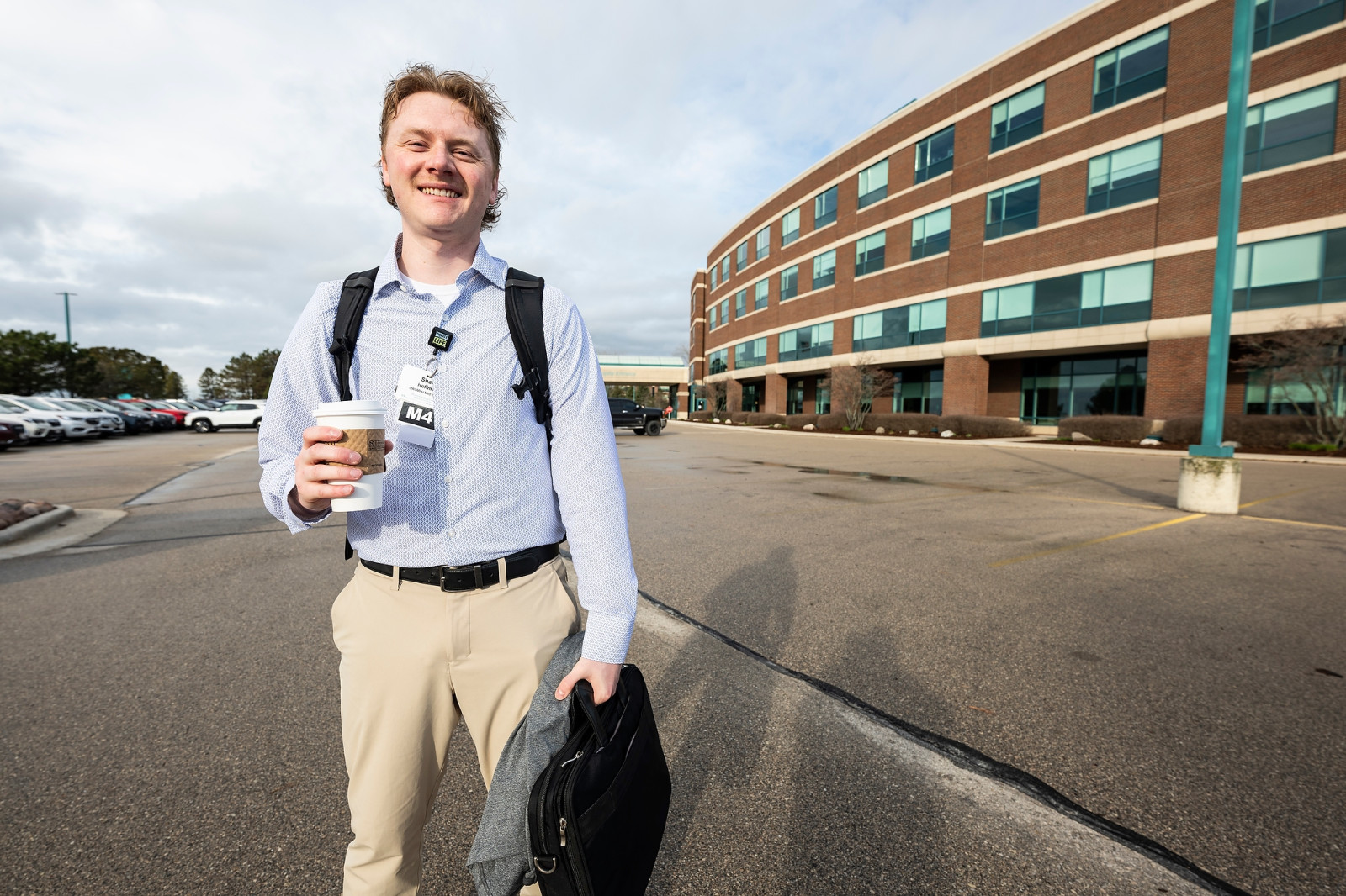 Hoffman holds a coffee up in one and a black briefcase in the other hand. He is standing in the parking lot of the Aurora Medical Center–Manitowoc County in Two Rivers. The medical center is pictured as a backdrop behind him.