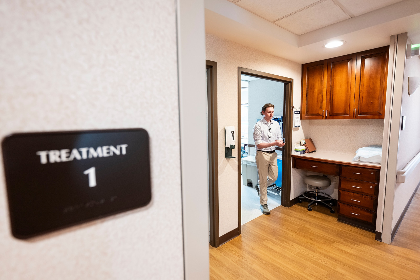 Hoffman walks out of a room in a medical clinic. As he exits, he's greeted by bright lights shining on the hallway and a built-in desk immediately outside of the treatment room.