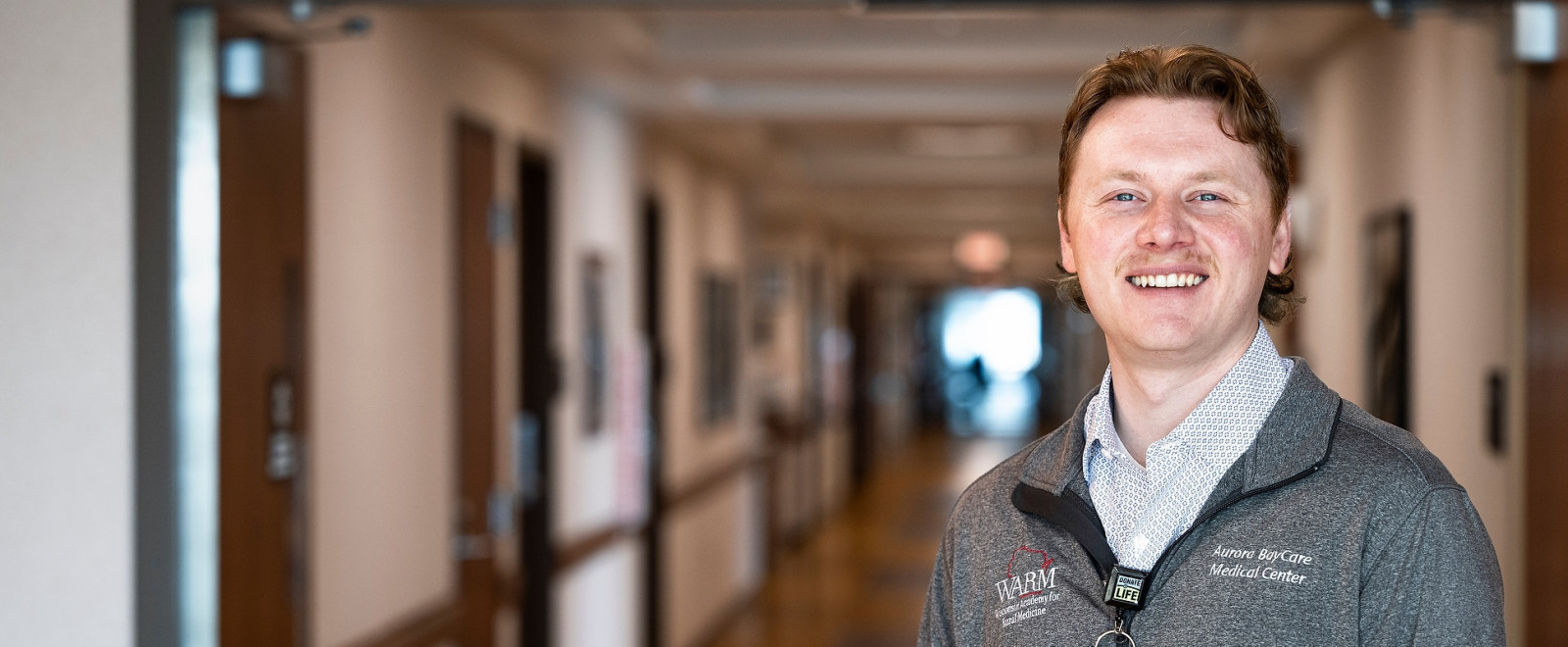 Shane stands in an empty hallway in the Aurora Medical Center–Manitowoc County in Two Rivers. The camera focuses on him as he smiles for a portrait.