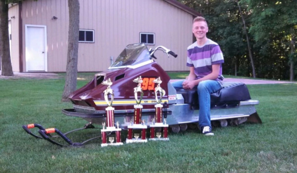 Hoffman sits atop a snowmobile parked in the green grass outside of a large shed. He smiles as three trophies are lined in front of the snowmobile.