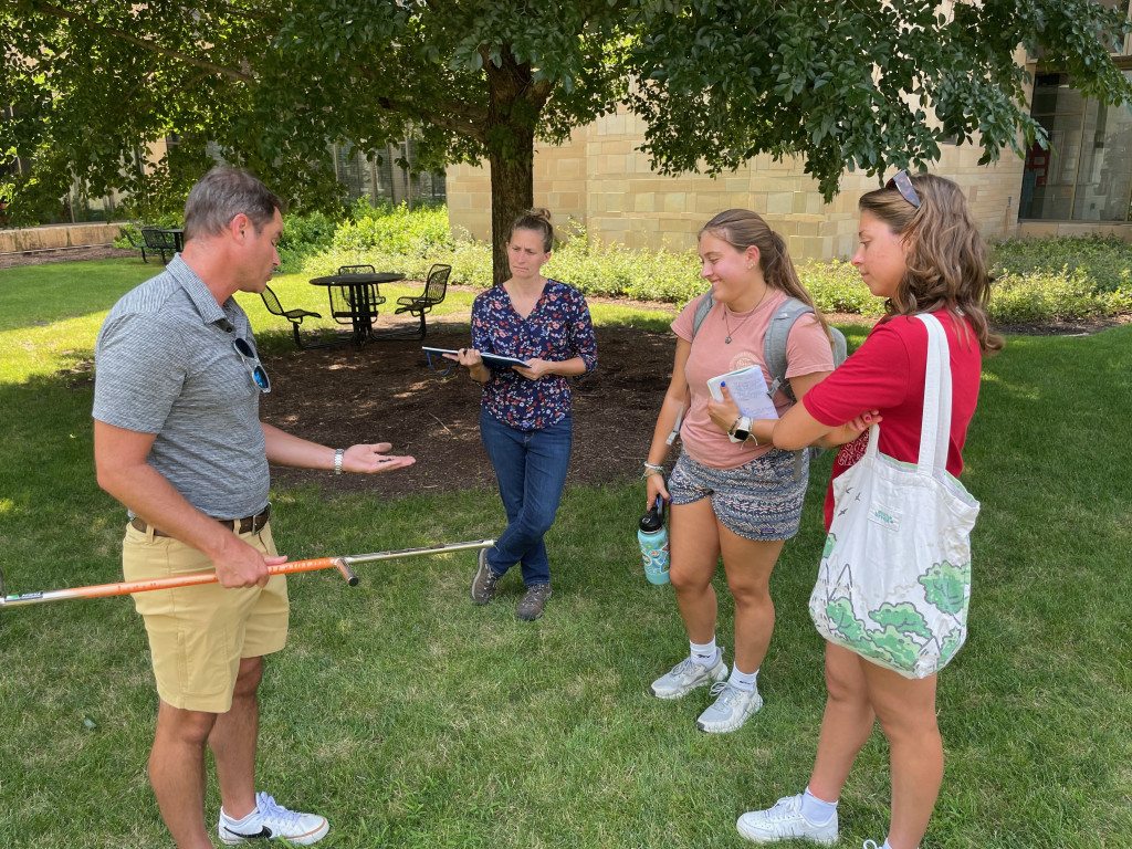 Several students and a man talk as they stand on a green lawn.