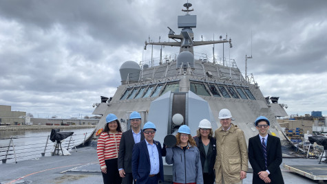 Five people in hard hats stand in a row and smile to the camera while standing on the deck of a U.S. Navy combat ship on an overcast day.
