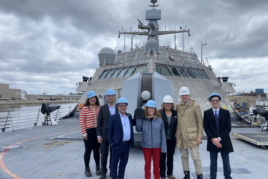 Five people in hard hats stand in a row and smile to the camera while standing on the deck of a U.S. Navy combat ship on an overcast day.