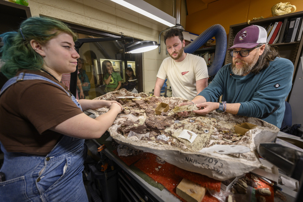 Three people stand around a table filled with fossils and rocks, and work carefully on them with tools.
