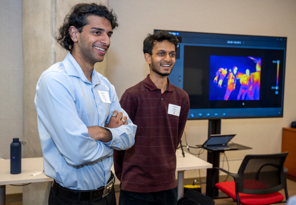 Tera Reddy and Basu stand in front of a table and a large TV screen that displays a still image of their project. They speak to event attendees who are not pictured in the photo.