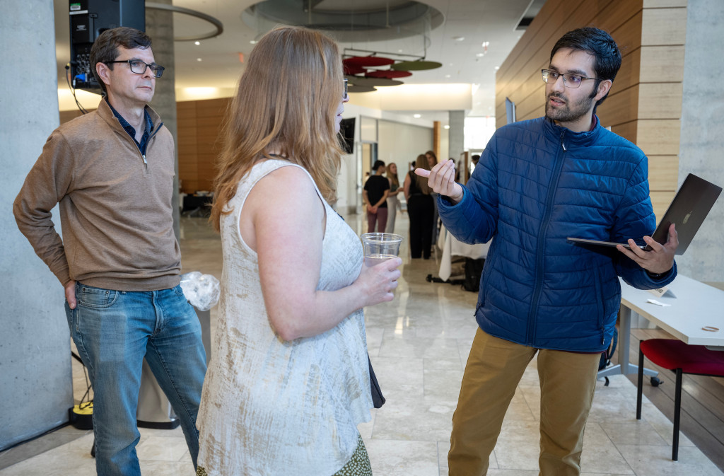 Vivek Deshpande stands with an open laptop in one arm. He is using his other hand to animatedly share his project idea to Hausman and another event attendee.