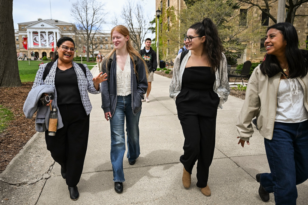 Four women walk along a sidewalk on a university campus, one of them talks and gestures as the others listen.