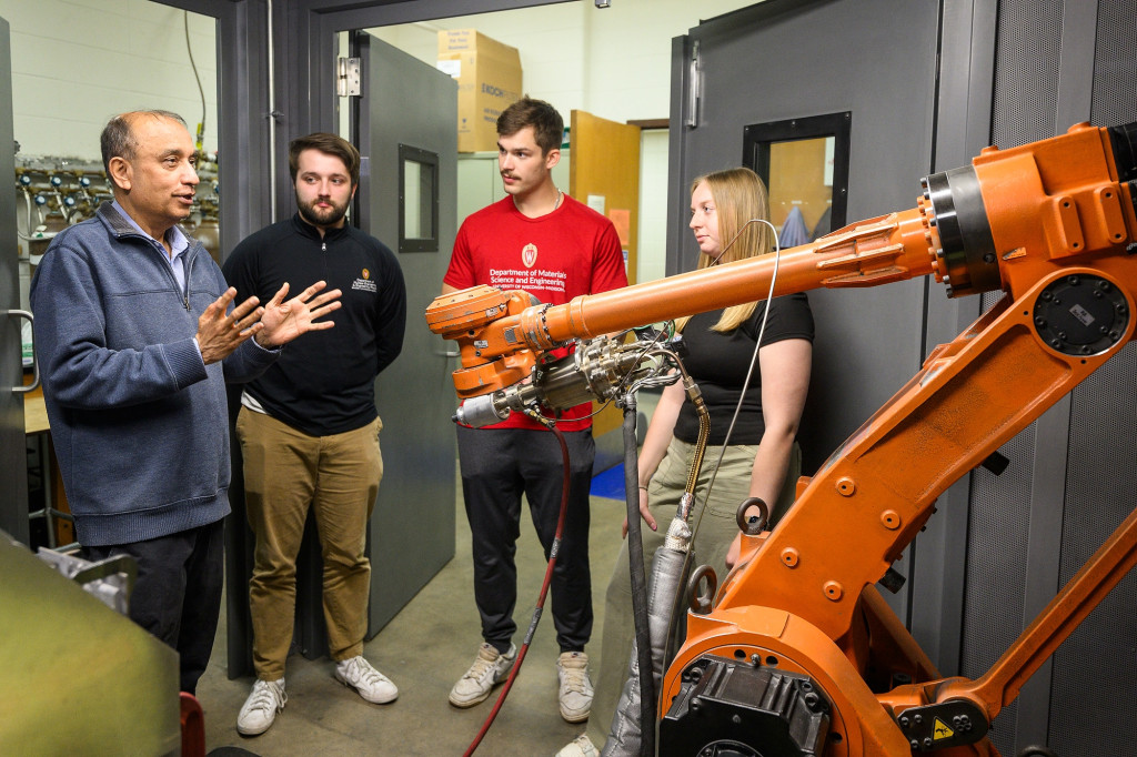 Four people are shown together in a laboratory with sophisticated electronic equipment. A man speaks and gestures as the other three listen.