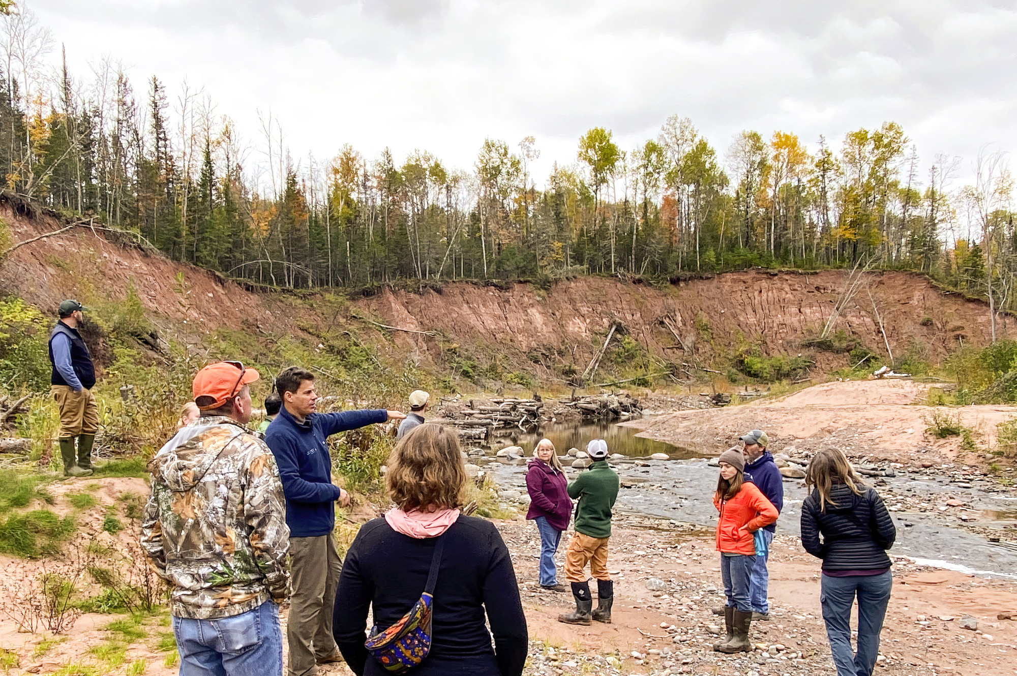 A group wearing jackets and boots stands along a forested bank that was caused by erosion.