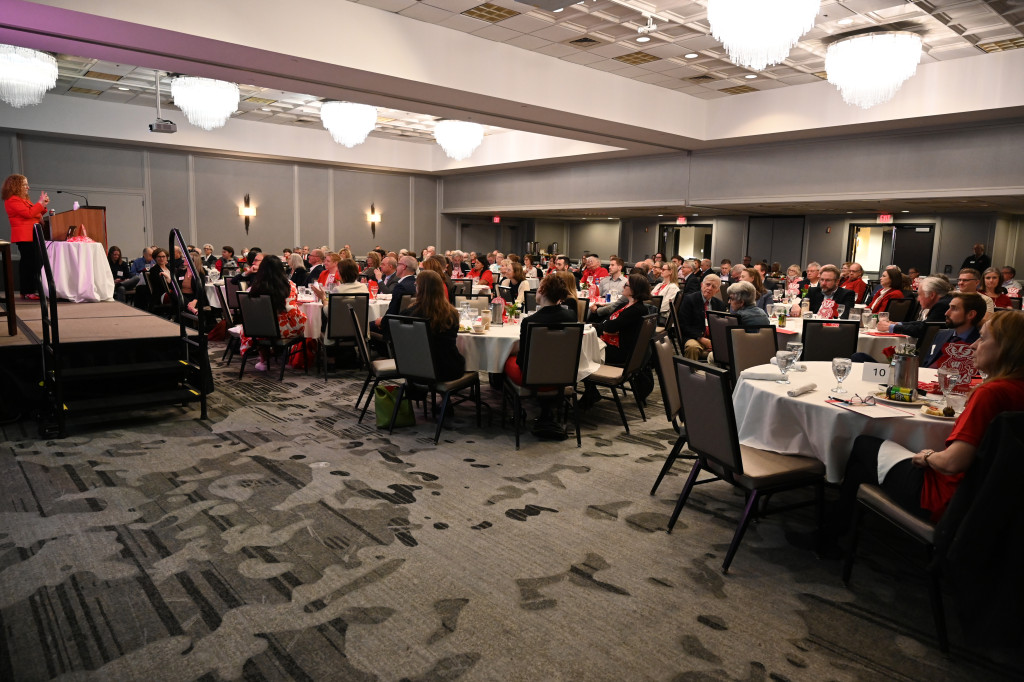 A carpeted room with brightly-lit chandeliers plays host to circular tables filled with alumni and key UW leaders. They face the stage on the left side of the image, where the Chancellor addresses them from behind a podium atop a small platform.