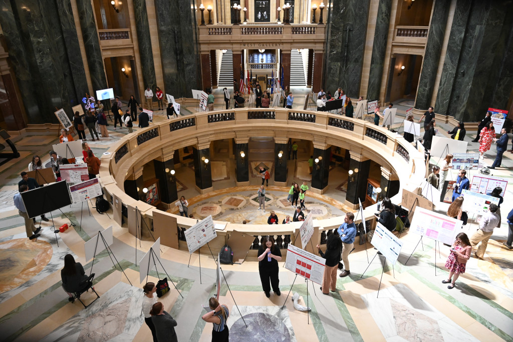 The Wisconsin State Capitol Rotunda is shown from above. UW-Madison researchers stand around the marble railing, each with designated tripod stand where their research project posters sit. The researchers speak with passersby about their work, which is summarized on the poster.
