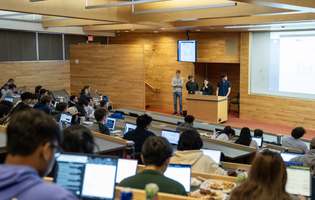 A team stands at the head of a room adorned with wood paneled walls. He gestures as he talks through his presentation to the crow d in front of him.