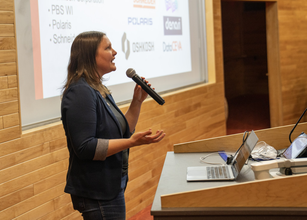 Feild stands behind a podium and addresses the filled classom using a microphone that she holds in one hand. 