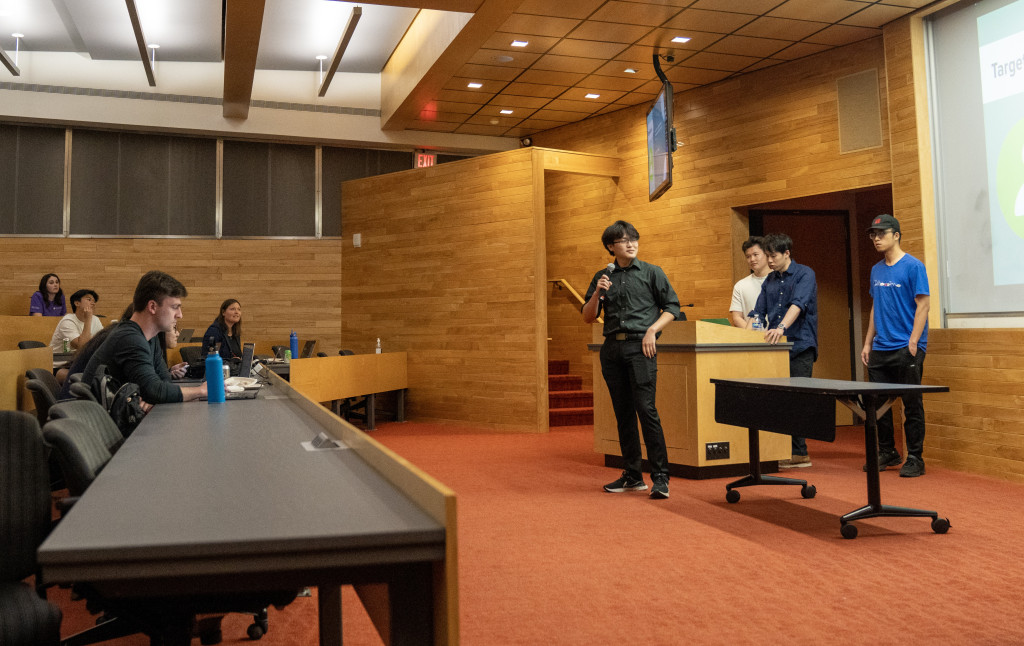 Zhu stands at the head of a room adorned with wood paneled walls. He gestures as he talks through his presentation to the crow d in front of him.