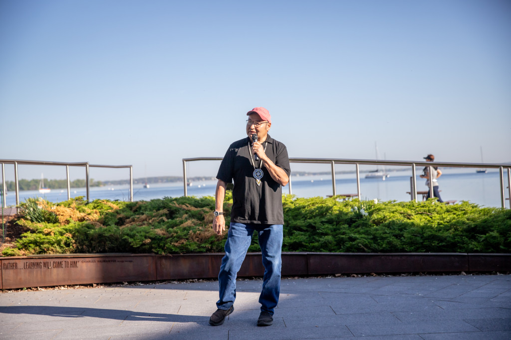 A man stands in front of a lake and talks into a microphone.