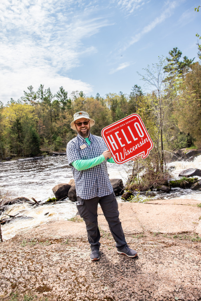 A man stands in front of a river with rapids in it, holding a sign reading