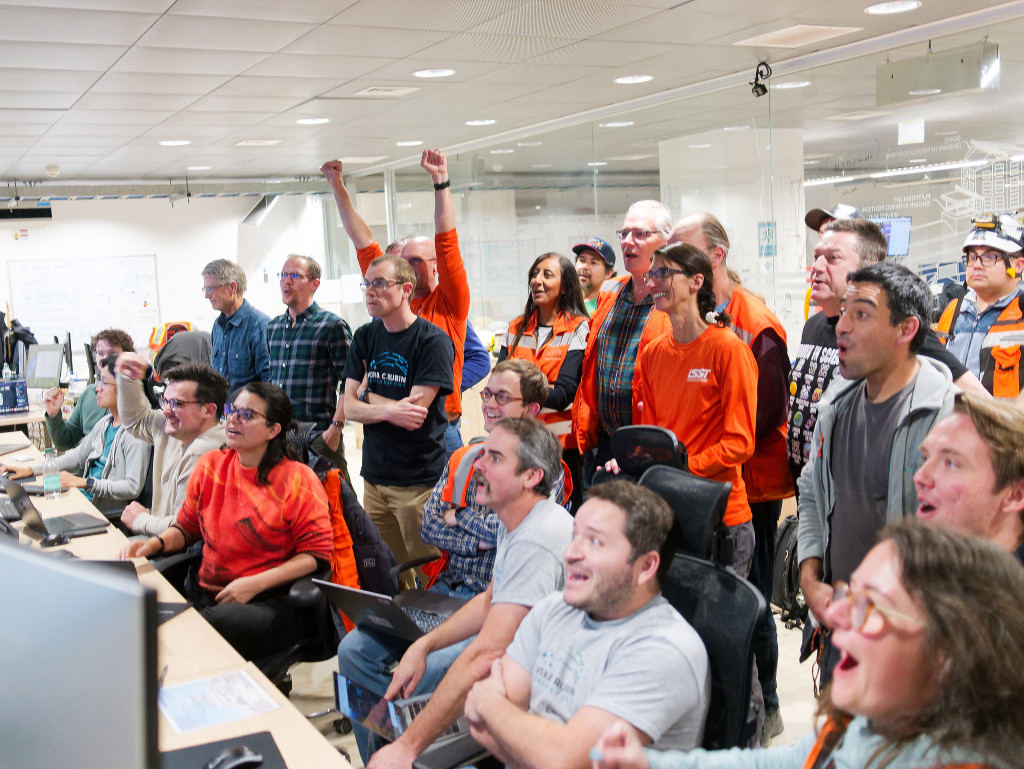 A group of astronauts, some sitting while others stand, watch in celebration and amazement as images produced by the telescope begin to broadcast on their screens.