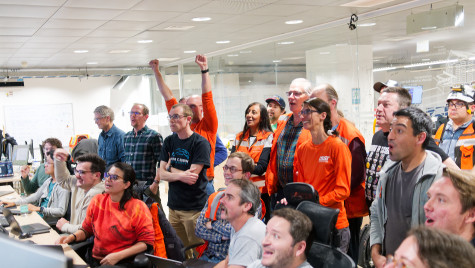 A group of astronauts, some sitting while others stand, watch in celebration and amazement as images produced by the telescope begin to broadcast on their screens.