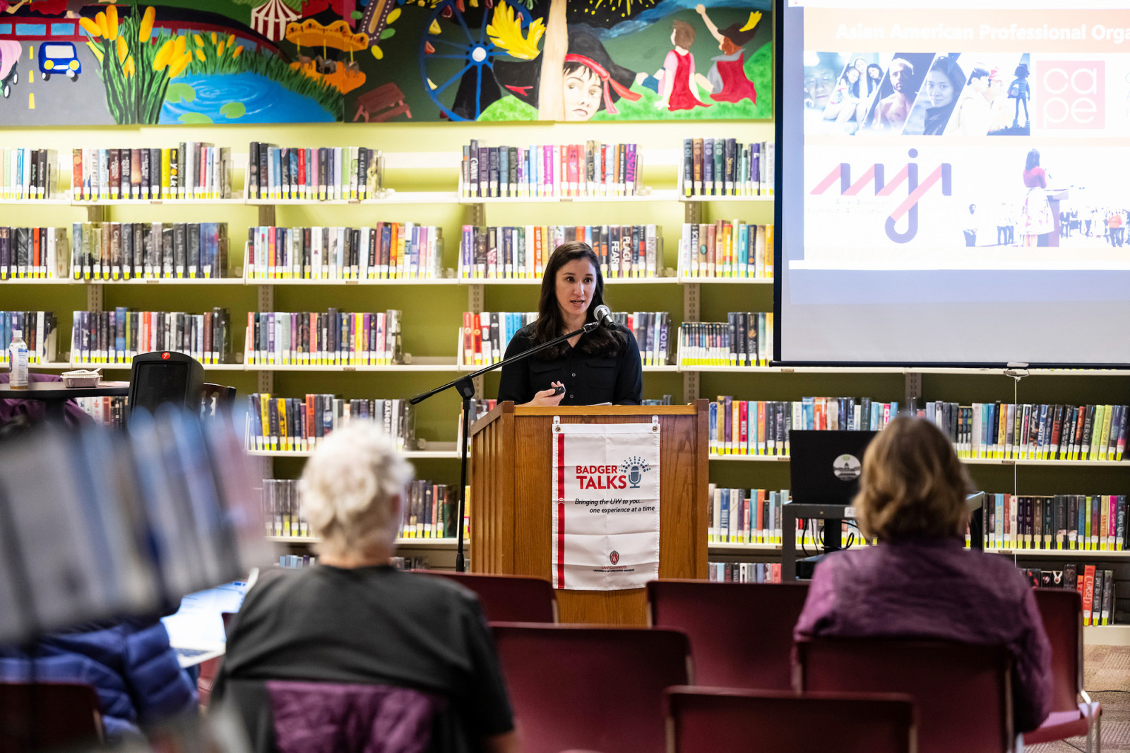 Lopez stands behind a podium with rows of books and display screen in the background. She speaks to seated viewers in the foreground.