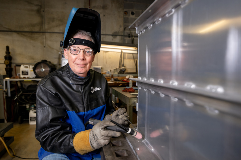 A man is shown wearing a welding mask turned upward and heavy gloves, by some equipment.