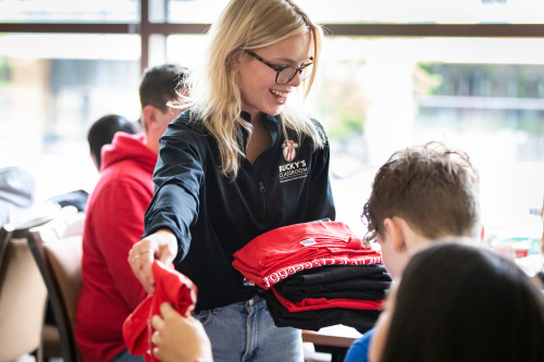A woman holds a stack of t-shirts and hands one off to a youth, as other kids surround her.