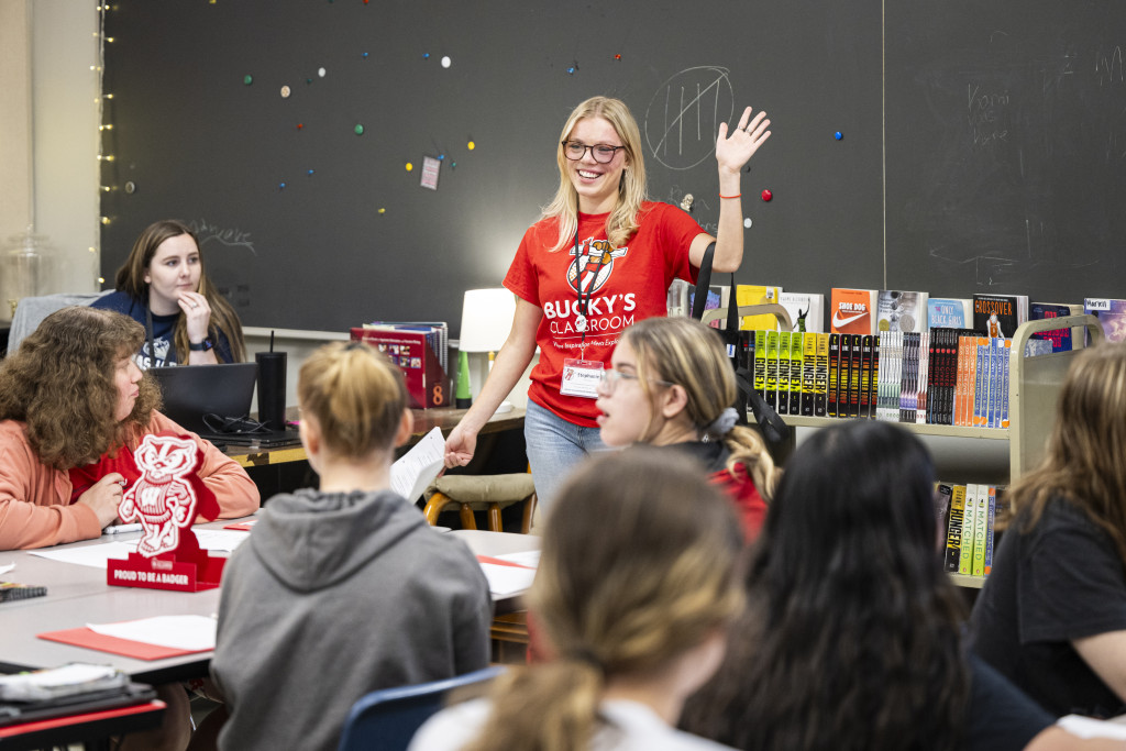 A woman in a red Bucky Badger shirt stands up in front of a class and talks and waves.