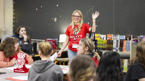 A woman in a red Bucky Badger shirt stands up in front of a class and talks and waves.
