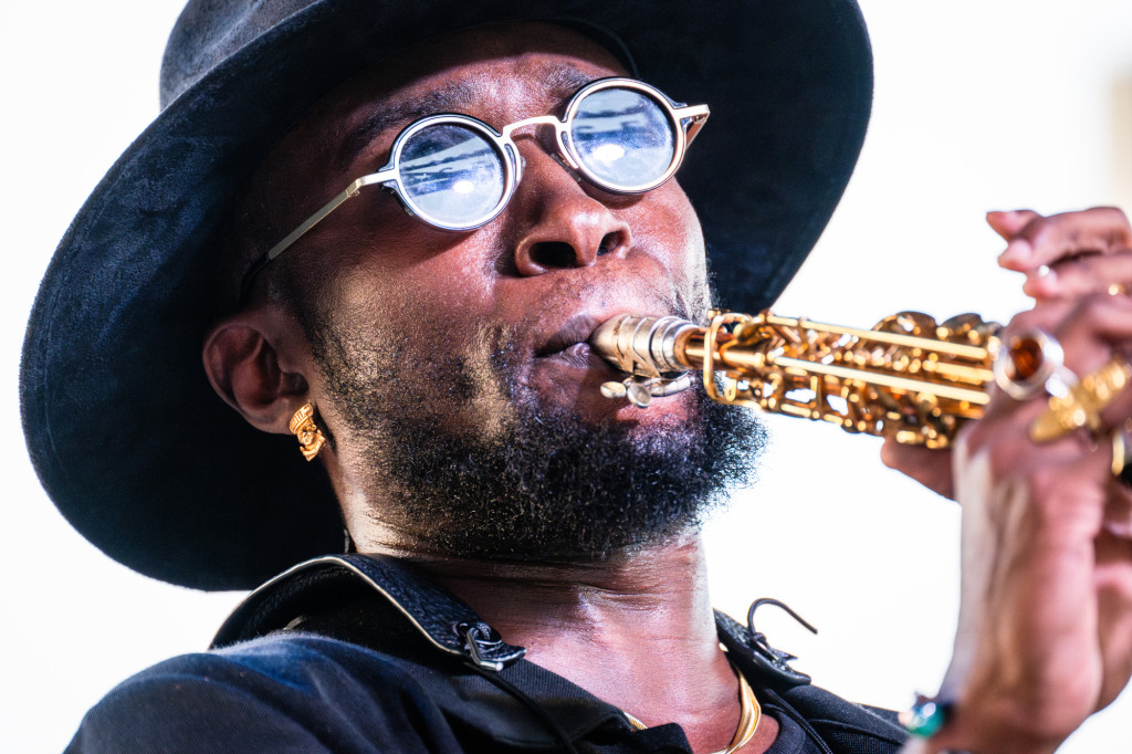 A man wearing sunglasses and hat blows into a saxophone.